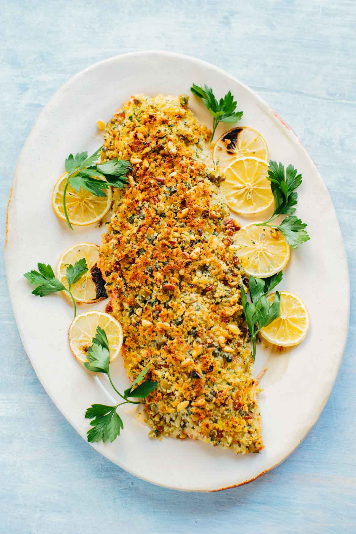 Overhead view of a complete salmon fillet on an oval white platter, topped with golden Sicilian breadcrumb mixture. Lemon slices and vibrant parsley sprigs artfully arranged around the fish enhance the Mediterranean presentation.
