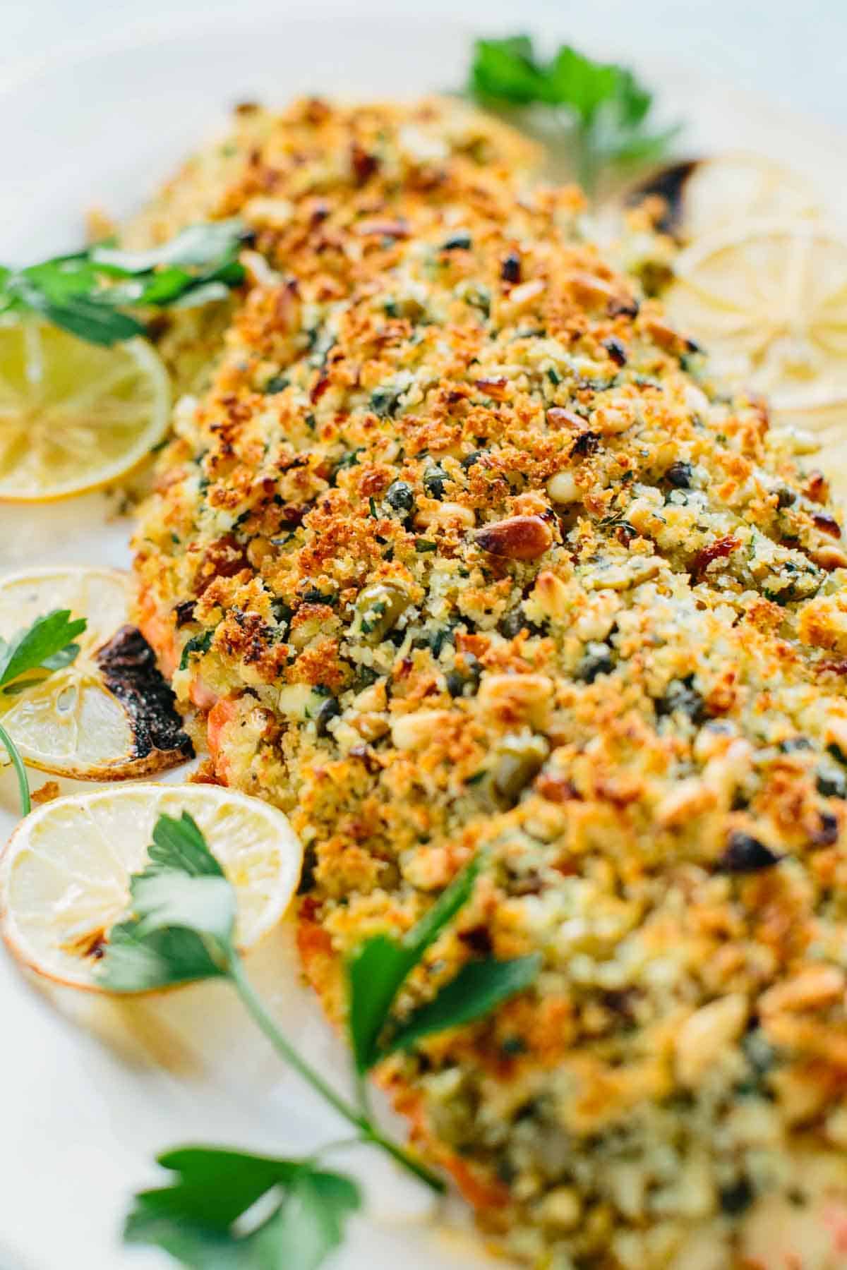 Close-up shot showing the detailed texture of the golden breadcrumb topping, revealing capers, pine nuts, and herbs embedded in the crust. Lemon wedges and parsley sprigs frame the perfectly baked salmon.