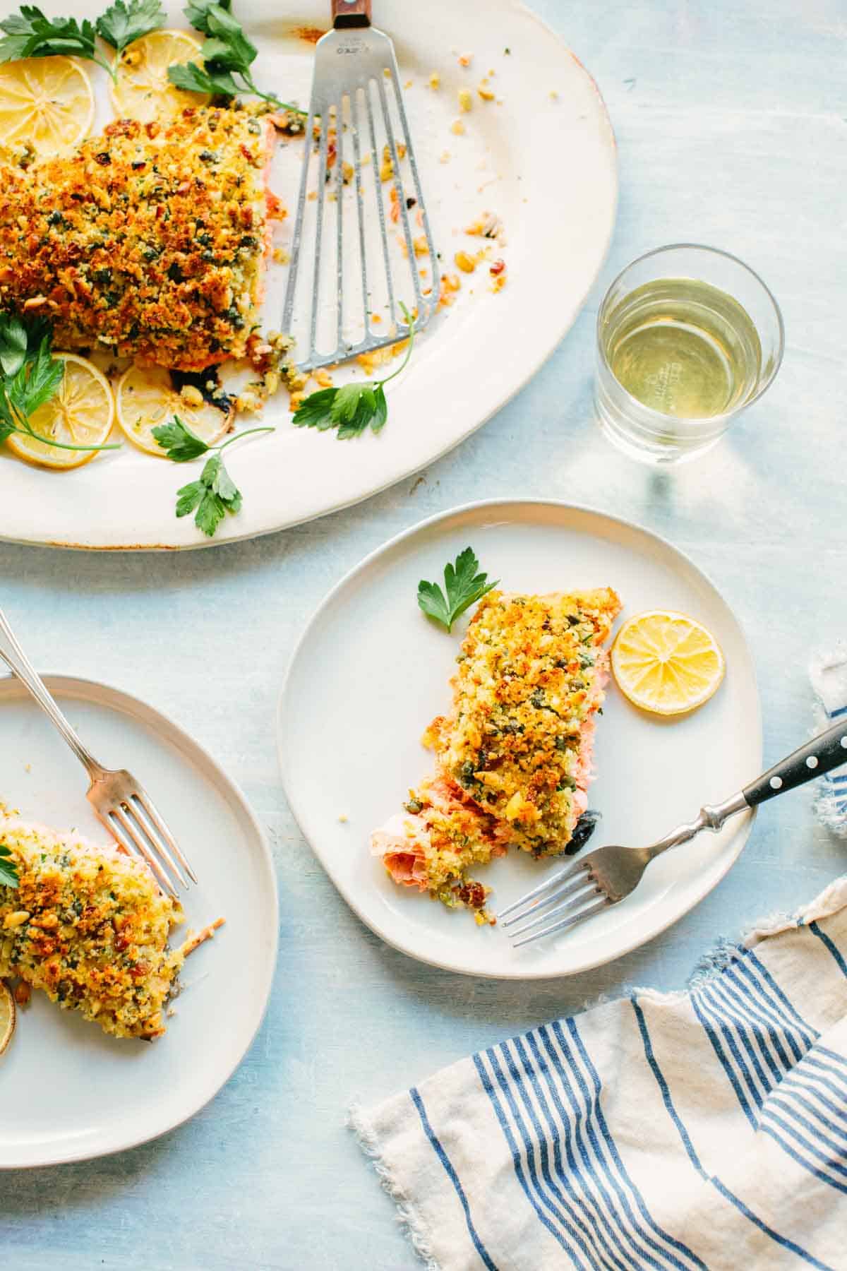Elegant table setting featuring the breadcrumb-topped salmon served family style alongside individual portions. A glass of white wine complements the meal, with blue-striped napkins adding a coastal touch.