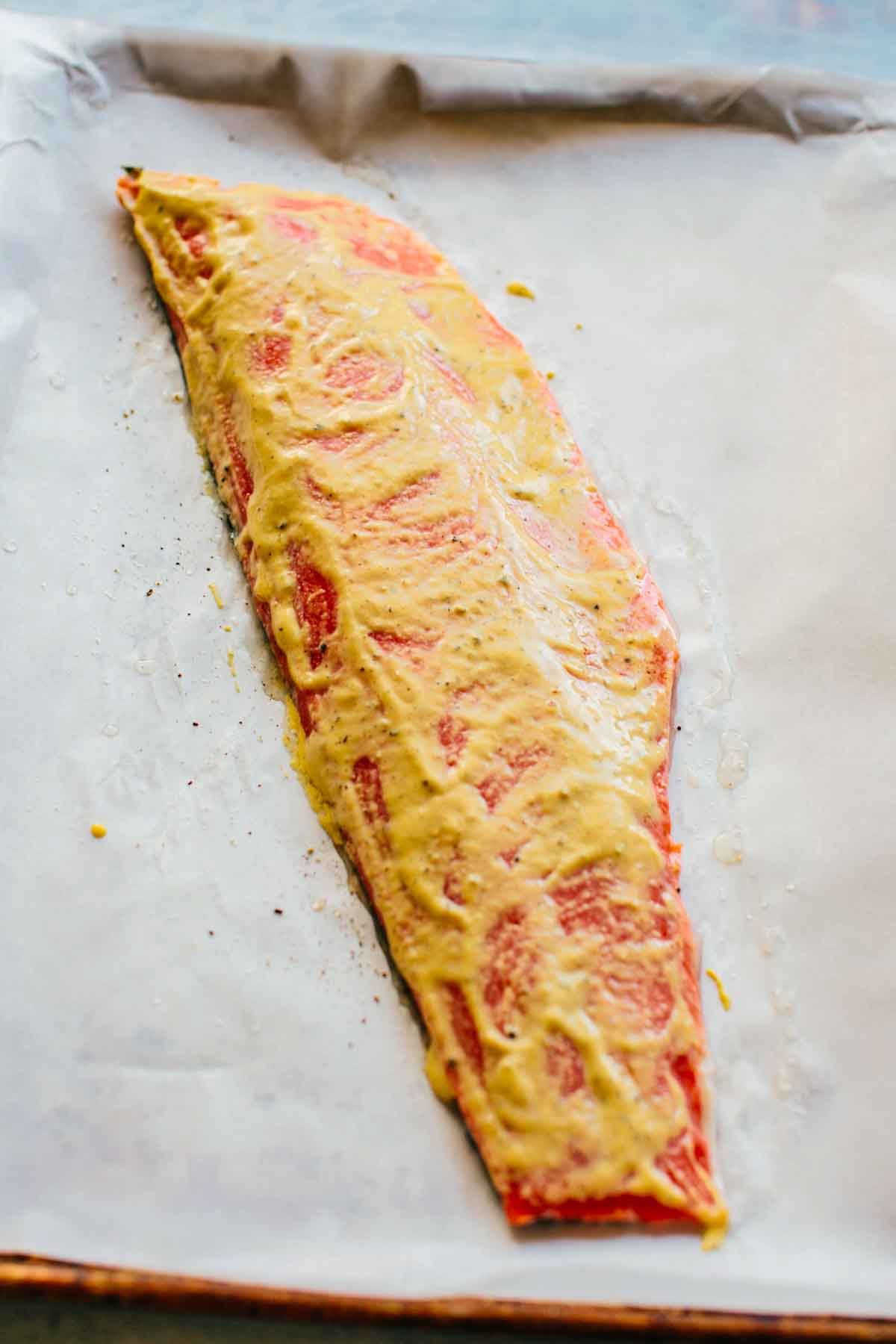 Raw salmon fillet brushed with a mustard-based binding mixture before baking, showing the preparation process. The vibrant pink fish on parchment paper is ready for the breadcrumb topping to be added.