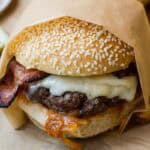 Close-up of a bacon cheddar burger wrapped in parchment, featuring a sesame seed bun, crispy bacon, melted cheese, and caramelized tomato onion jam.
