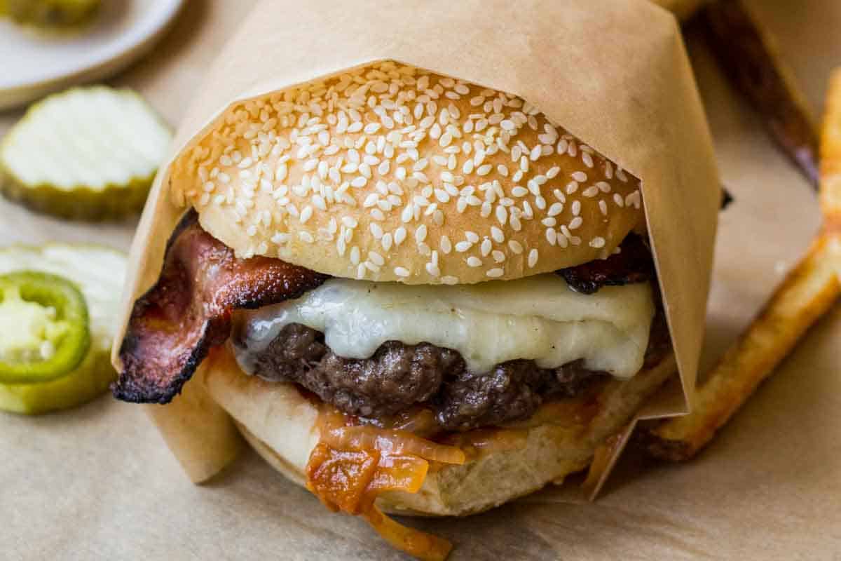 Close-up of a bacon cheddar burger wrapped in parchment, featuring a sesame seed bun, crispy bacon, melted cheese, and caramelized tomato onion jam.