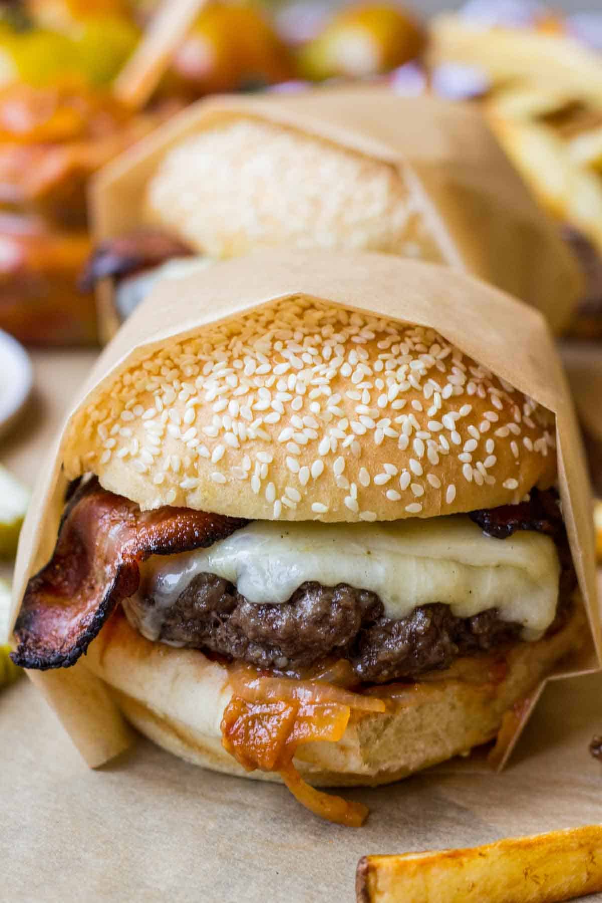 Front-facing view of a sesame seed bun burger with melted cheddar, bacon, and tomato onion jam, wrapped in parchment paper with fries and pickles on the side.
