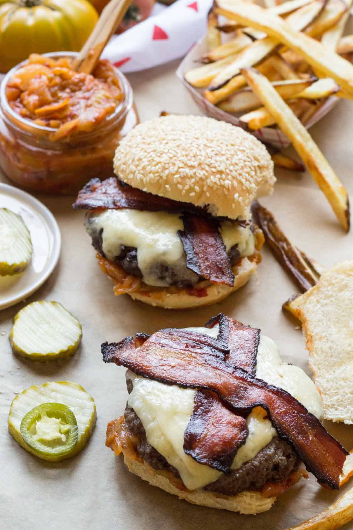 Overhead view of two open bacon cheddar burgers on parchment with pickles, fries, and a jar of tomato onion jam in the background.