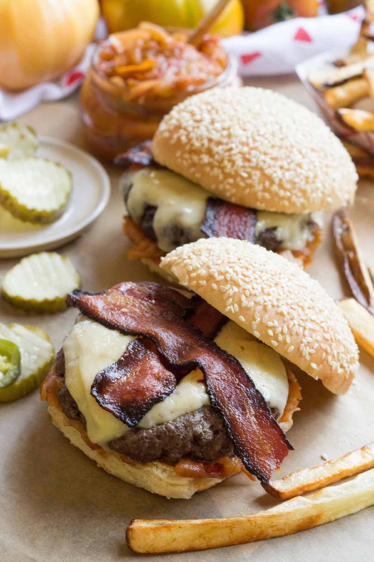 Two assembled bacon cheddar burgers with sesame buns and crispy bacon, served on parchment with a side of fries and pickles.