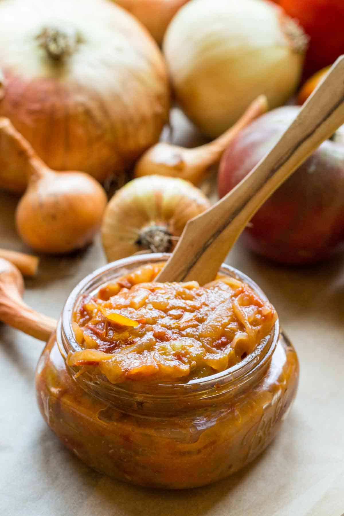 Small jar of chunky tomato onion jam with a wooden spoon, surrounded by heirloom onions and tomatoes on a rustic surface.