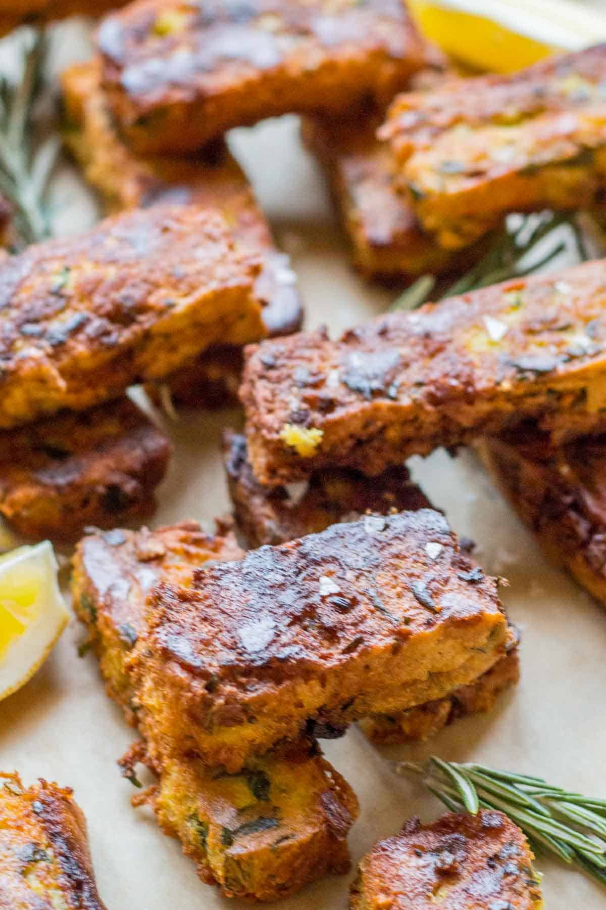 Close-up of golden-brown chickpea veggie fritters arranged on a light surface. The rectangularly-shaped fritters show a crispy exterior with flecks of herbs and vegetables visible within. Fresh rosemary sprigs and lemon wedges garnish the platter, adding aromatic and citrus accents.