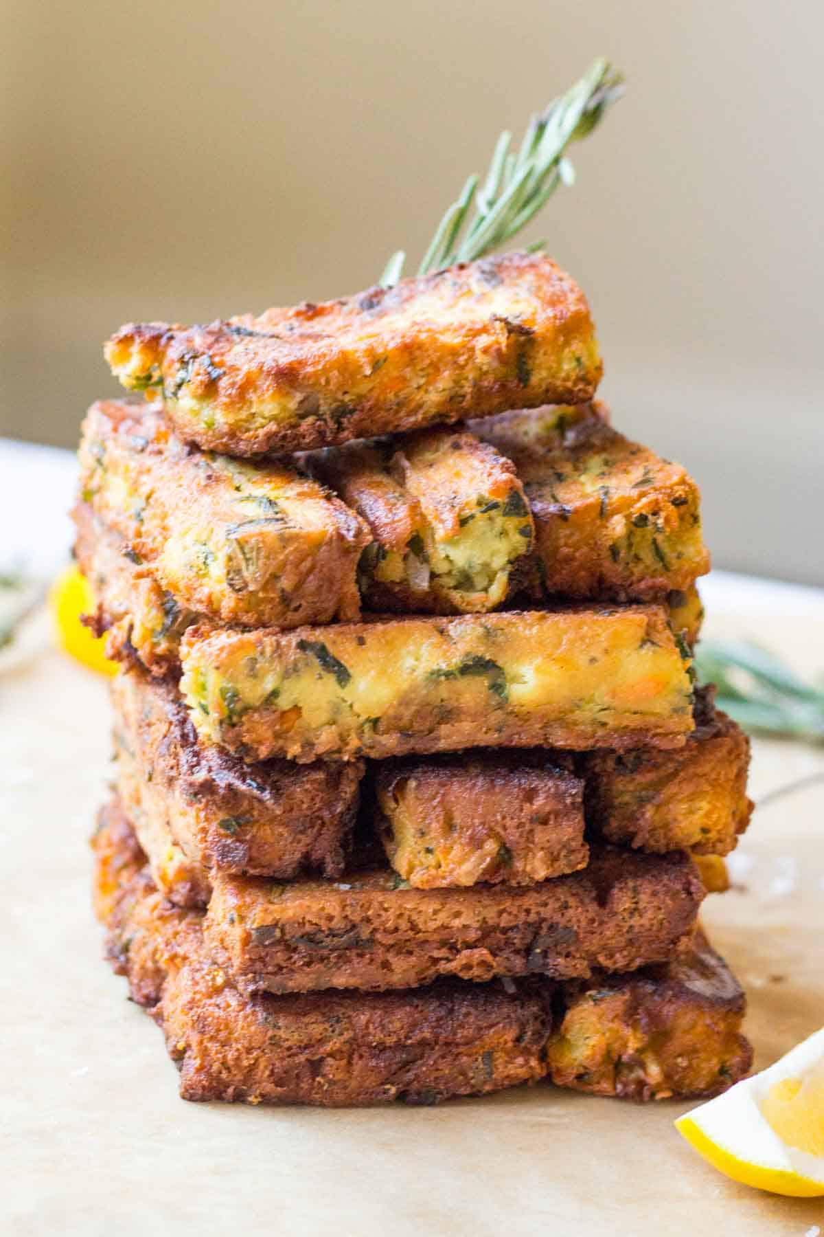 A tall stack of chickpea veggie fritters arranged in a cross-hatch pattern, showcasing their sturdy texture. The golden-brown fritters are flecked with green herbs and have a beautifully caramelized exterior. A sprig of rosemary tops the stack, with a lemon wedge visible at the bottom of the frame.