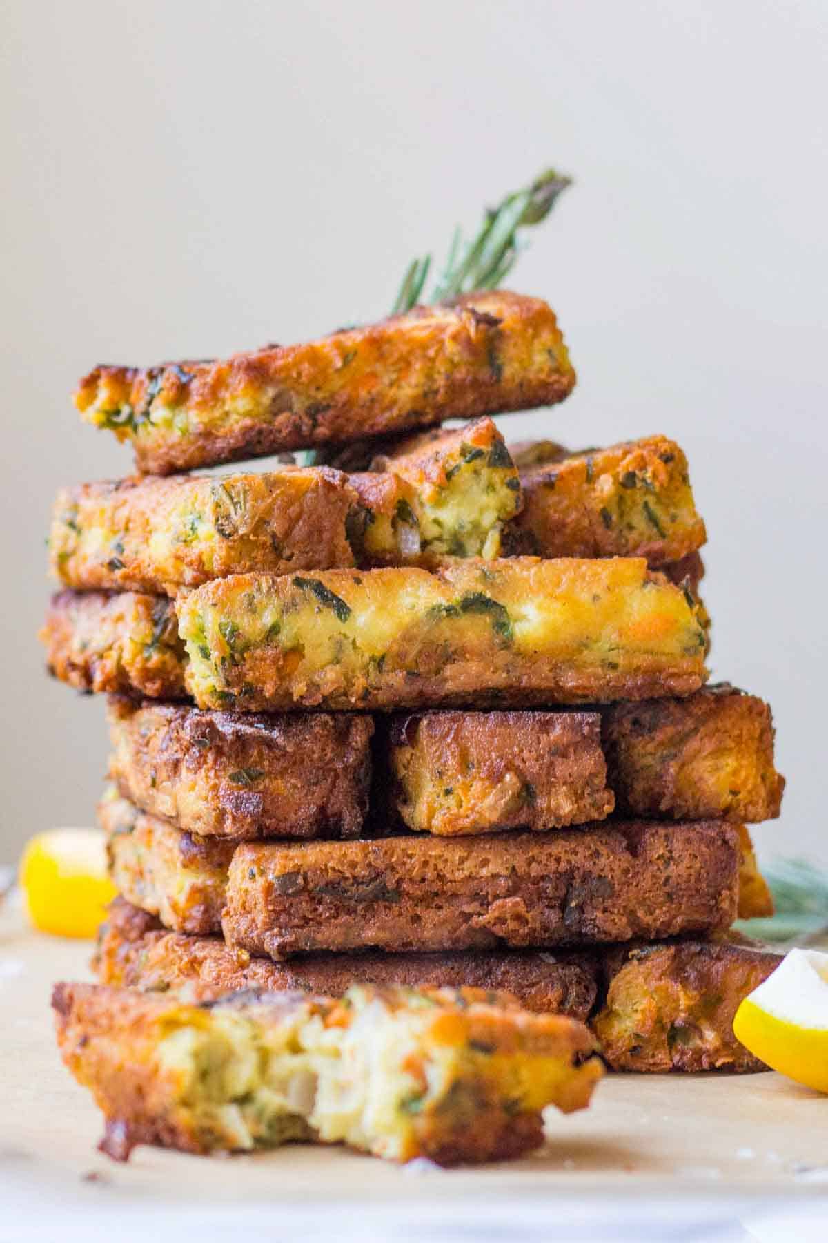A tall stack of rectangular chickpea fritters with a slightly different arrangement. The cross-section of a fritter is visible at the bottom, revealing the moist interior studded with vegetables and herbs, contrasting with the crispy exterior. Lemon wedges frame the composition.