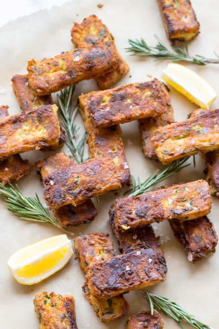 Overhead view of chickpea fritters arranged on parchment paper. The rectangular fritters are garnished with fresh rosemary sprigs and lemon wedges. Their golden-brown surface shows visible herbs and seasoning, with specks of salt visible on the crispy crust. The arrangement appears casual yet appetizing.