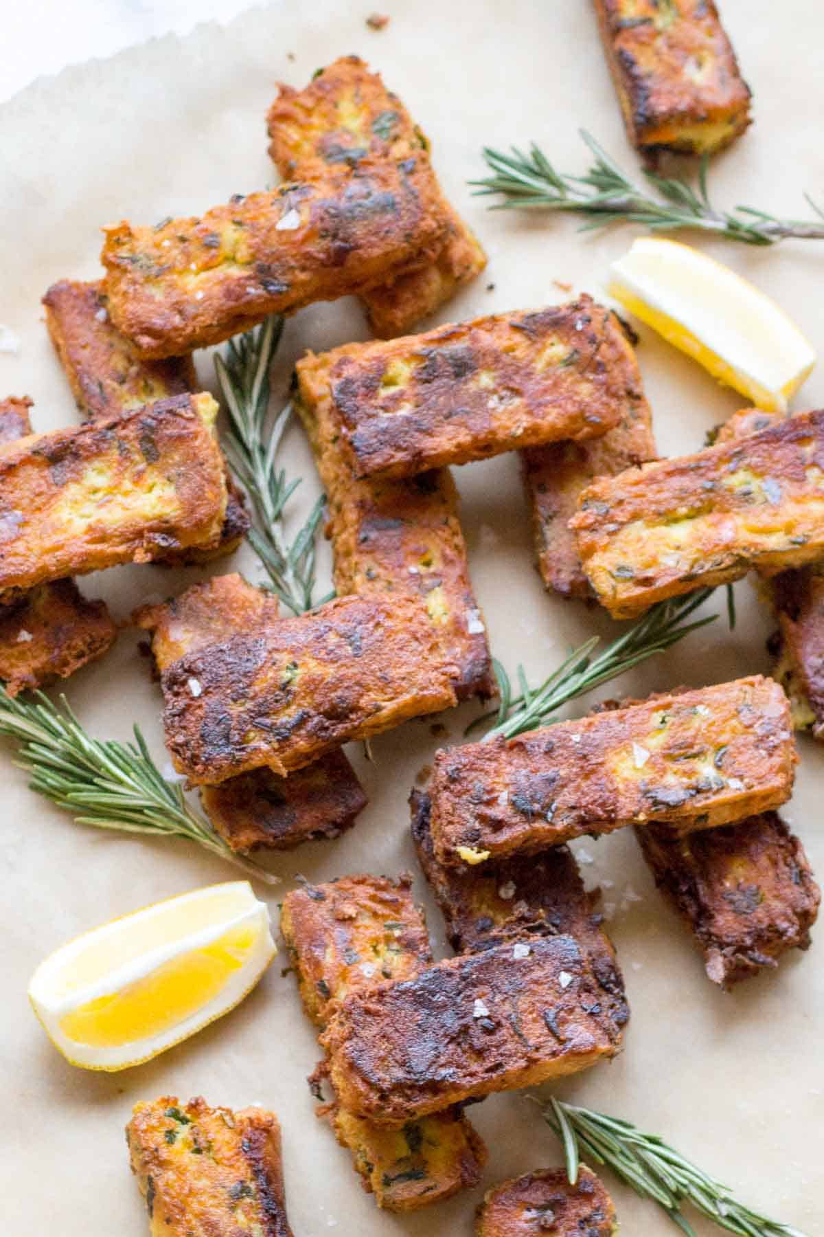 Overhead view of chickpea fritters arranged on parchment paper. The rectangular fritters are garnished with fresh rosemary sprigs and lemon wedges. Their golden-brown surface shows visible herbs and seasoning, with specks of salt visible on the crispy crust. The arrangement appears casual yet appetizing.
