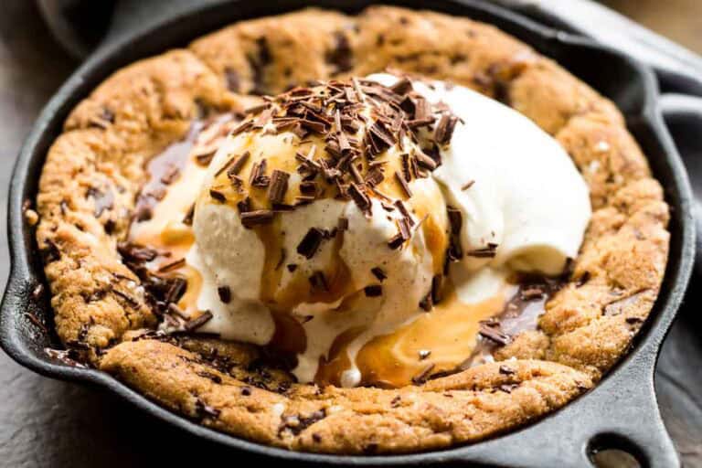 Close-up of a freshly baked chocolate chip cookie in a cast iron skillet topped with vanilla ice cream, chocolate shavings, and caramel sauce. The warm cookie edges are golden-brown and crisp while the center remains soft beneath the melting ice cream.