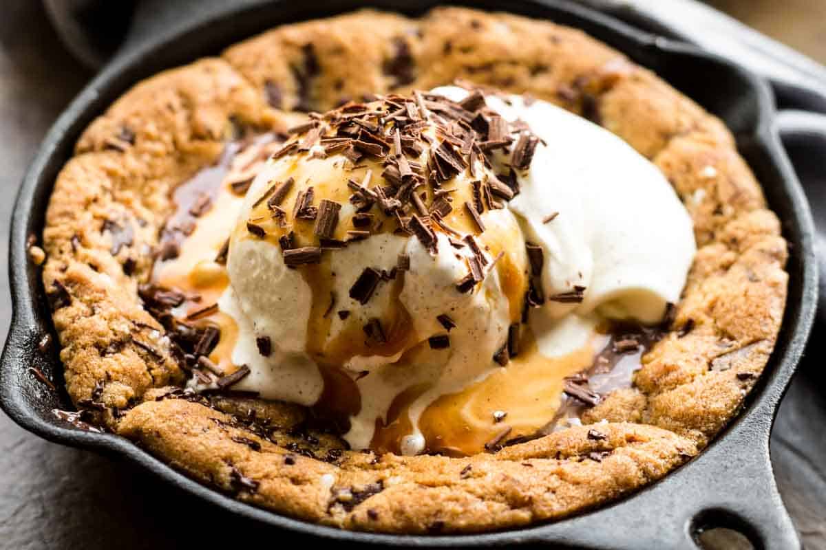 Close-up of a freshly baked chocolate chip cookie in a cast iron skillet topped with vanilla ice cream, chocolate shavings, and caramel sauce. The warm cookie edges are golden-brown and crisp while the center remains soft beneath the melting ice cream.