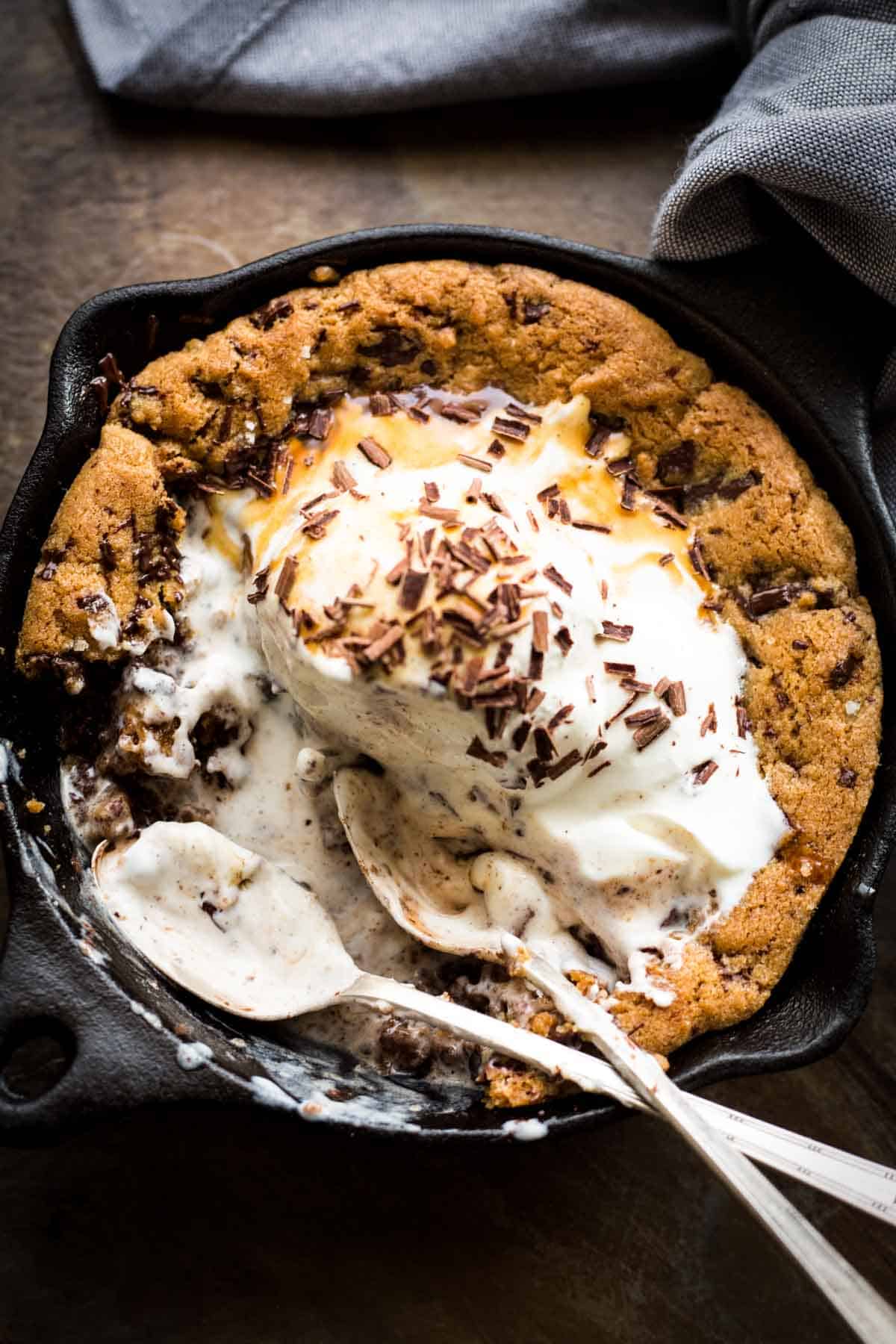 Partially eaten skillet cookie with vanilla ice cream and chocolate shavings, showing the gooey interior where the chocolate chips have melted. A spoon rests in the skillet, scooping through the dessert's soft center against the crisp cookie edge.