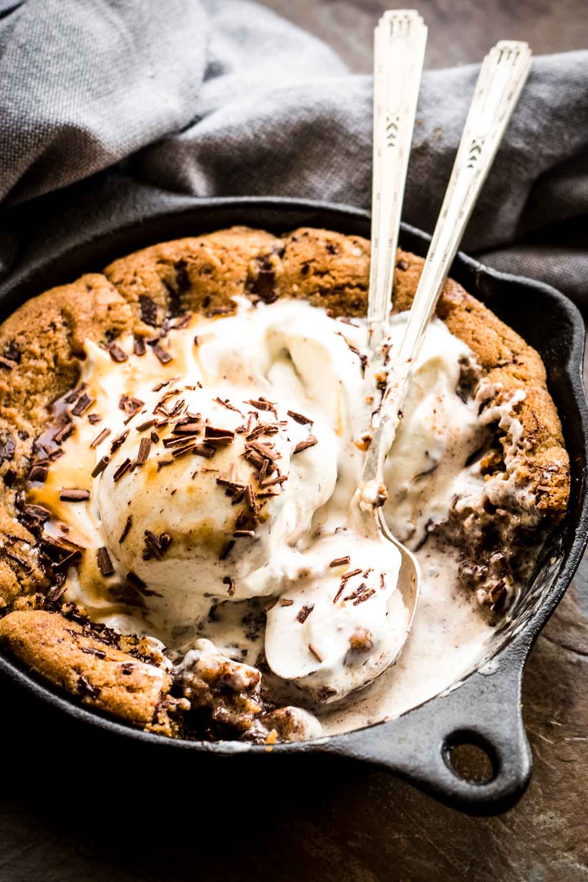 Artistic angle of a skillet cookie topped with melting vanilla ice cream, chocolate shavings, and caramel drizzle. Two vintage spoons stand upright in the dessert, ready for sharing, while the cookie begins to soften beneath the warm-cold contrast.