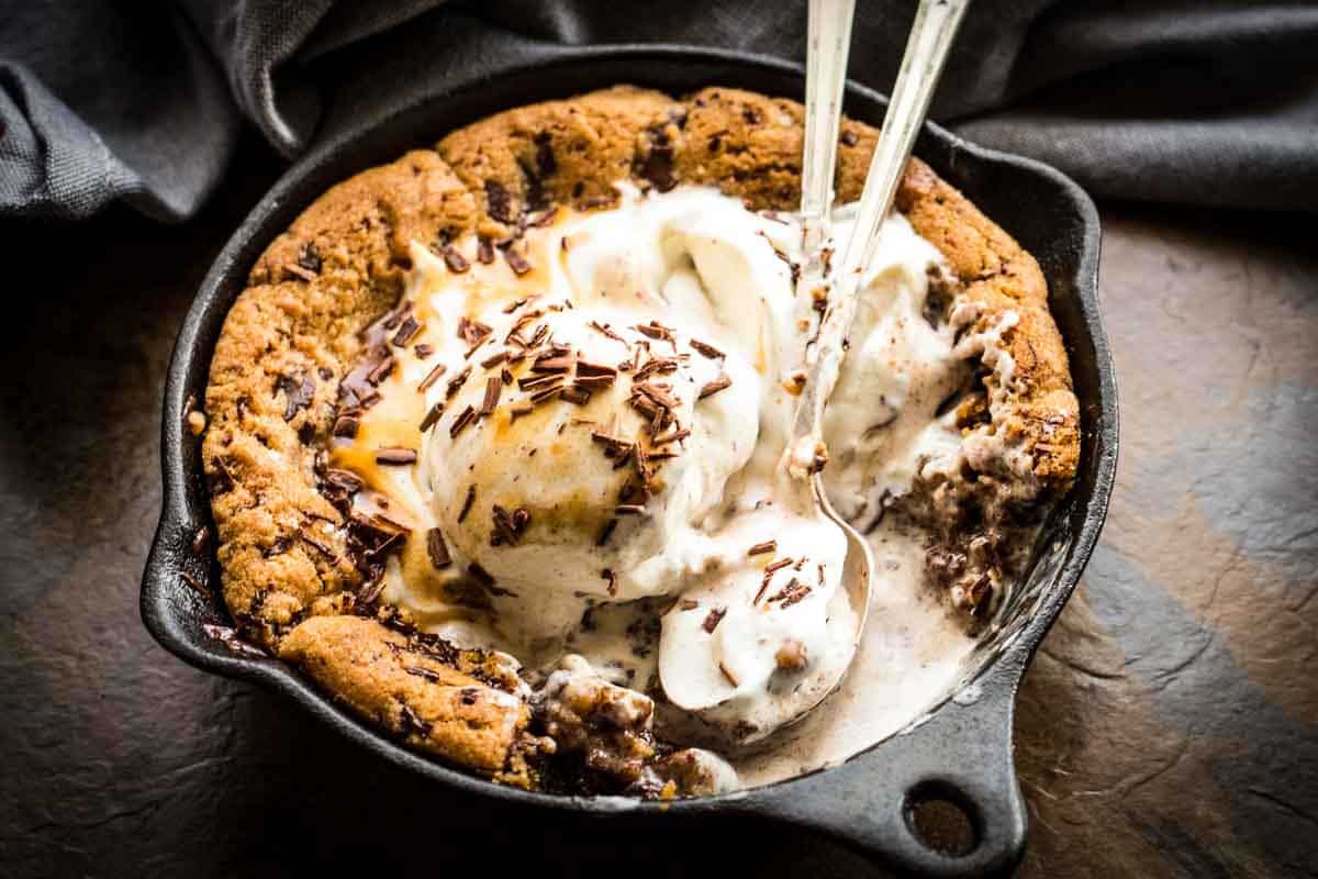 Similar angle to Image 3 showing the skillet cookie with vanilla ice cream and chocolate shavings against a dark background. The cast iron skillet creates a rustic presentation while highlighting the cookie's perfect golden exterior and melting toppings.