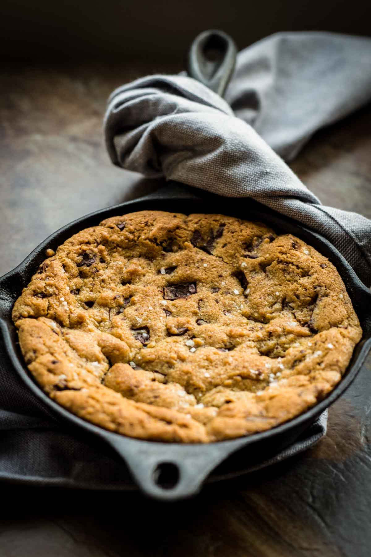 Freshly baked skillet chocolate chip cookie before adding toppings, displaying its perfectly golden surface studded with chocolate chunks and a light sprinkle of sea salt. A gray linen napkin frames the cast iron pan on a dark wooden surface.