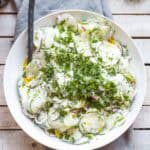 Overhead view of a white bowl filled with cucumber radish salad in a creamy yogurt dressing, garnished with fresh dill, set on a rustic wood surface with cucumbers and herbs.