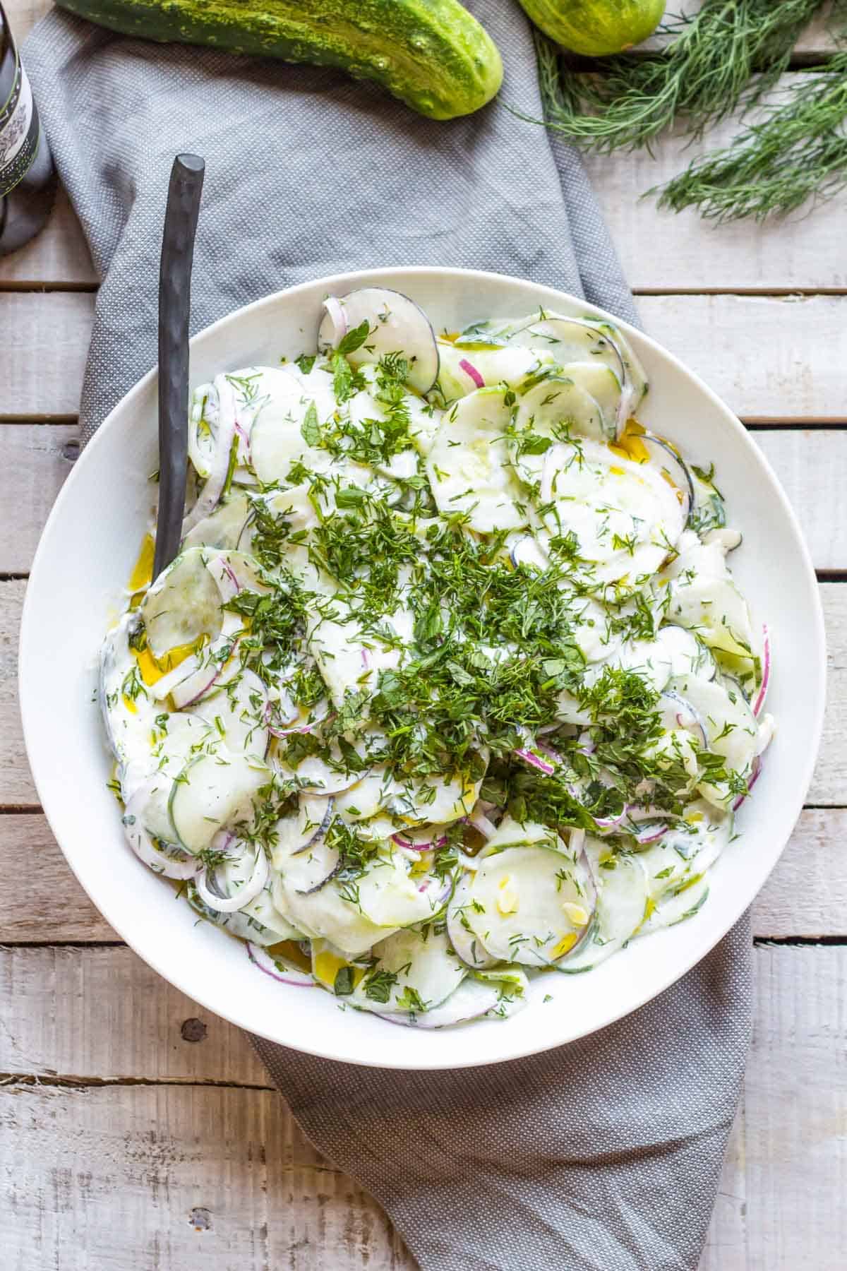 Overhead view of a white bowl filled with cucumber radish salad in a creamy yogurt dressing, garnished with fresh dill, set on a rustic wood surface with cucumbers and herbs.
