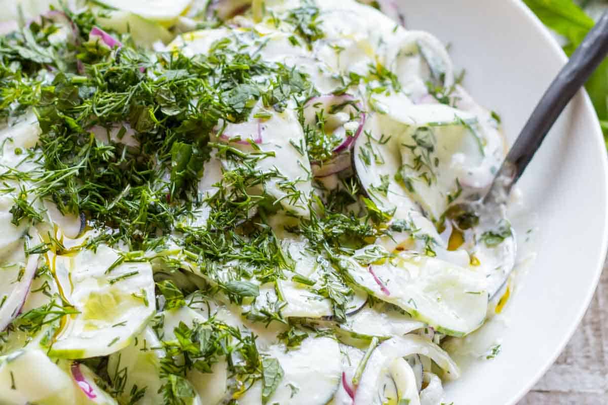 Close-up of a creamy cucumber and radish salad with yogurt dressing, red onions, and chopped fresh herbs, topped generously with dill and mint.