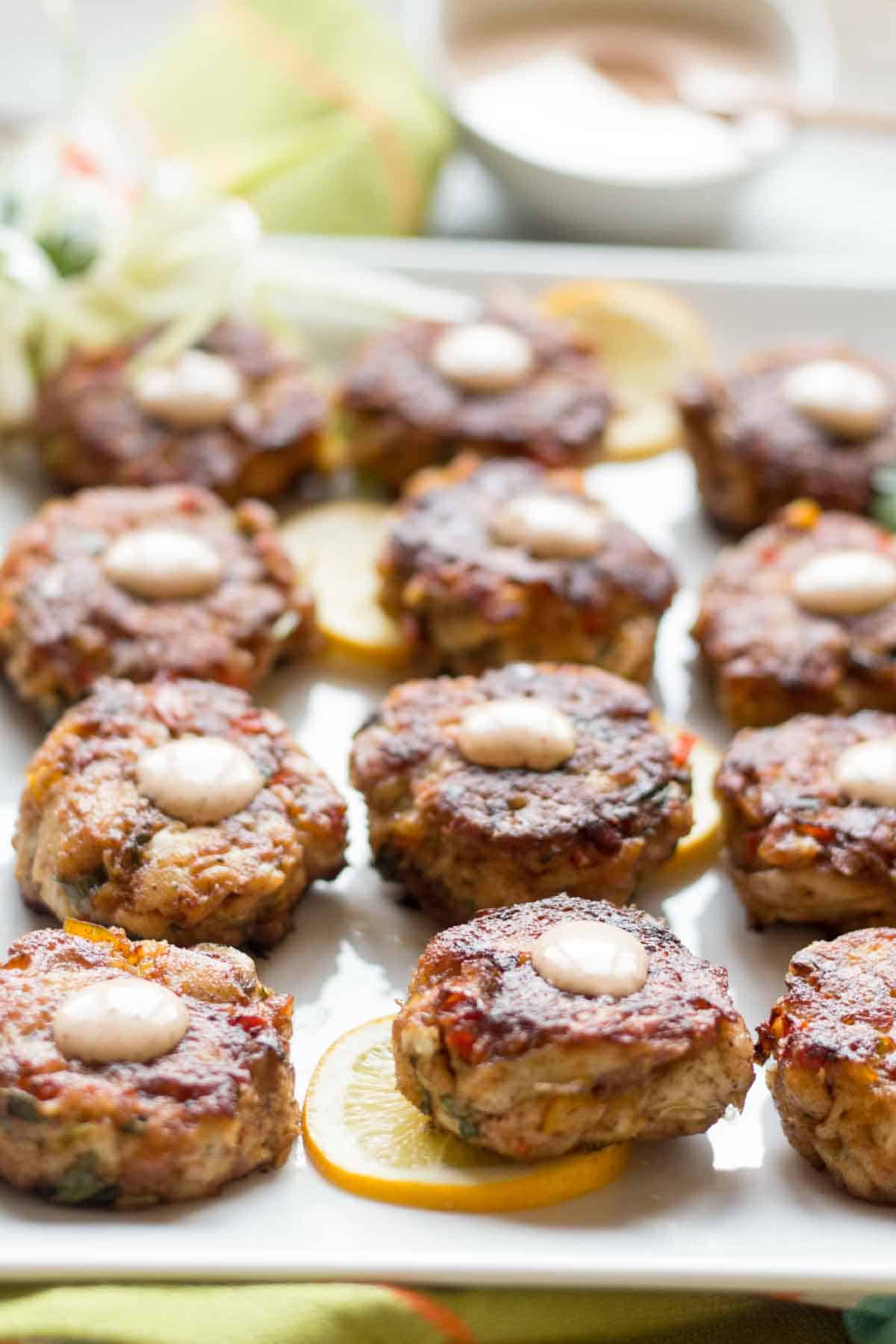 Close-up view of perfectly seared fish cakes on a white serving plate, showcasing their crispy exterior and flecked interior of seafood and herbs. Each cake features a uniform creamy topping and sits atop lemon slices.