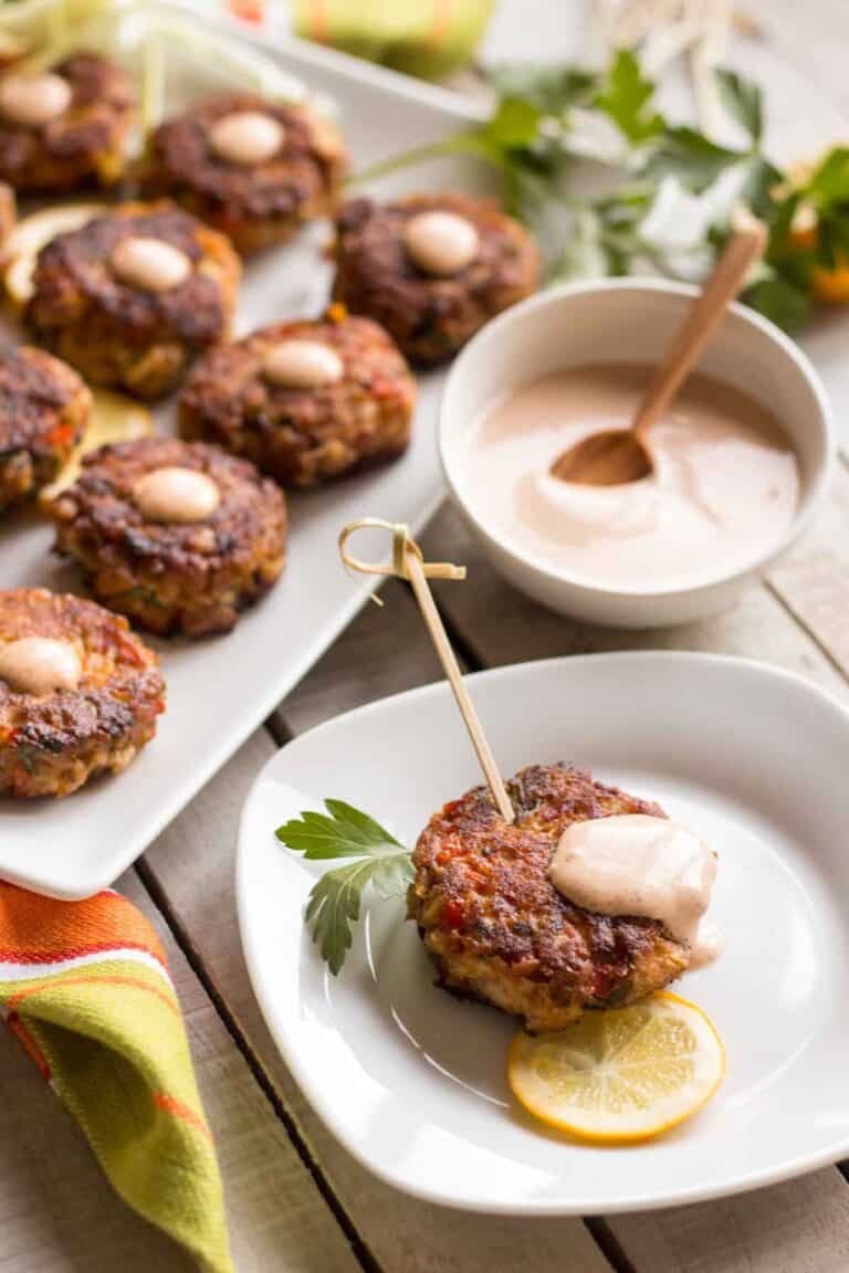 Elegant serving setup with a platter of fish cakes alongside individual portions and dipping sauce. One fish cake speared with a wooden pick sits on a small plate with a lemon slice, while a small bowl of creamy sauce with wooden spoon accompanies the arrangement.