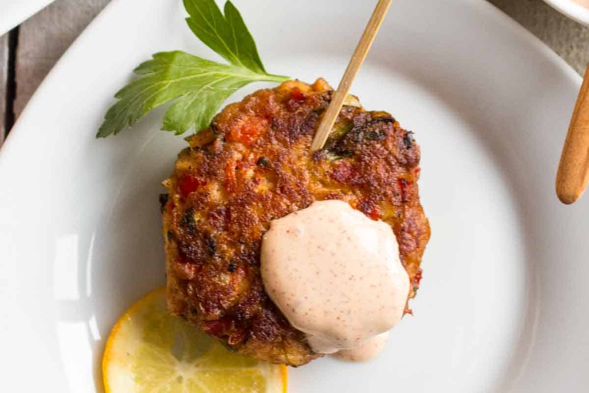 Detailed overhead shot of a single fish cake on a white plate, highlighting its golden-brown crust, visible flakes of fish and colorful vegetable pieces. A wooden toothpick and fresh parsley garnish the cake, which is topped with creamy sauce.