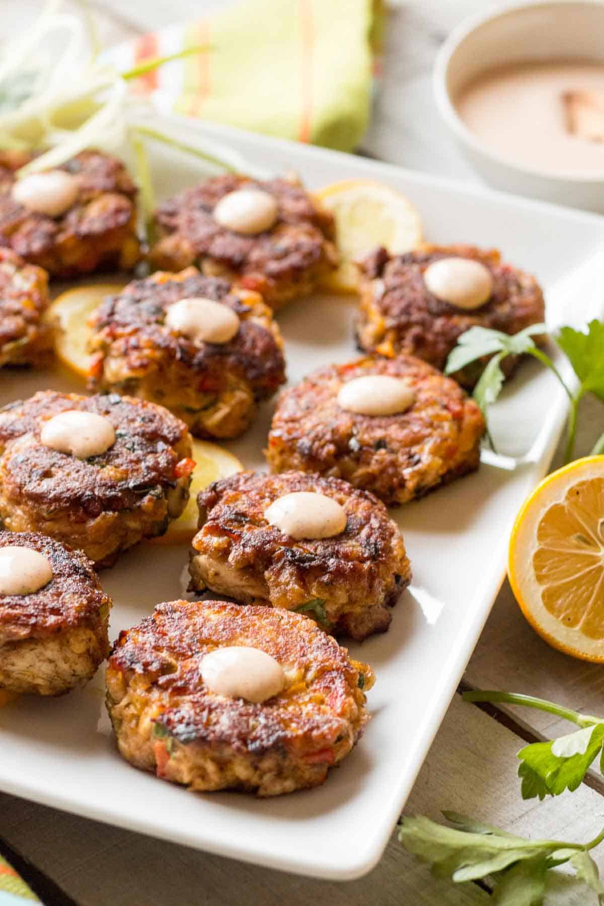 Angled view of fish cakes arranged in rows on a rectangular platter, showing their uniform size and consistent browning. Fresh herbs and lemon wedges adorn the plate, with a small dish of dipping sauce visible in the background.