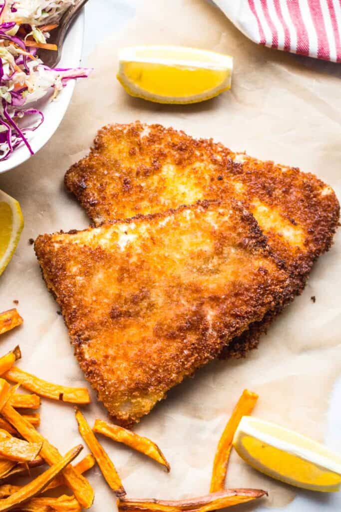 An overhead view of golden-brown, triangular panko-crusted fish fillets on parchment paper. The crispy coating has a rich amber color with darker spots where it's most deeply fried. Lemon wedges are placed around the fish, with sweet potato fries visible at the bottom of the frame and a small portion of coleslaw with purple cabbage visible in the upper left corner.