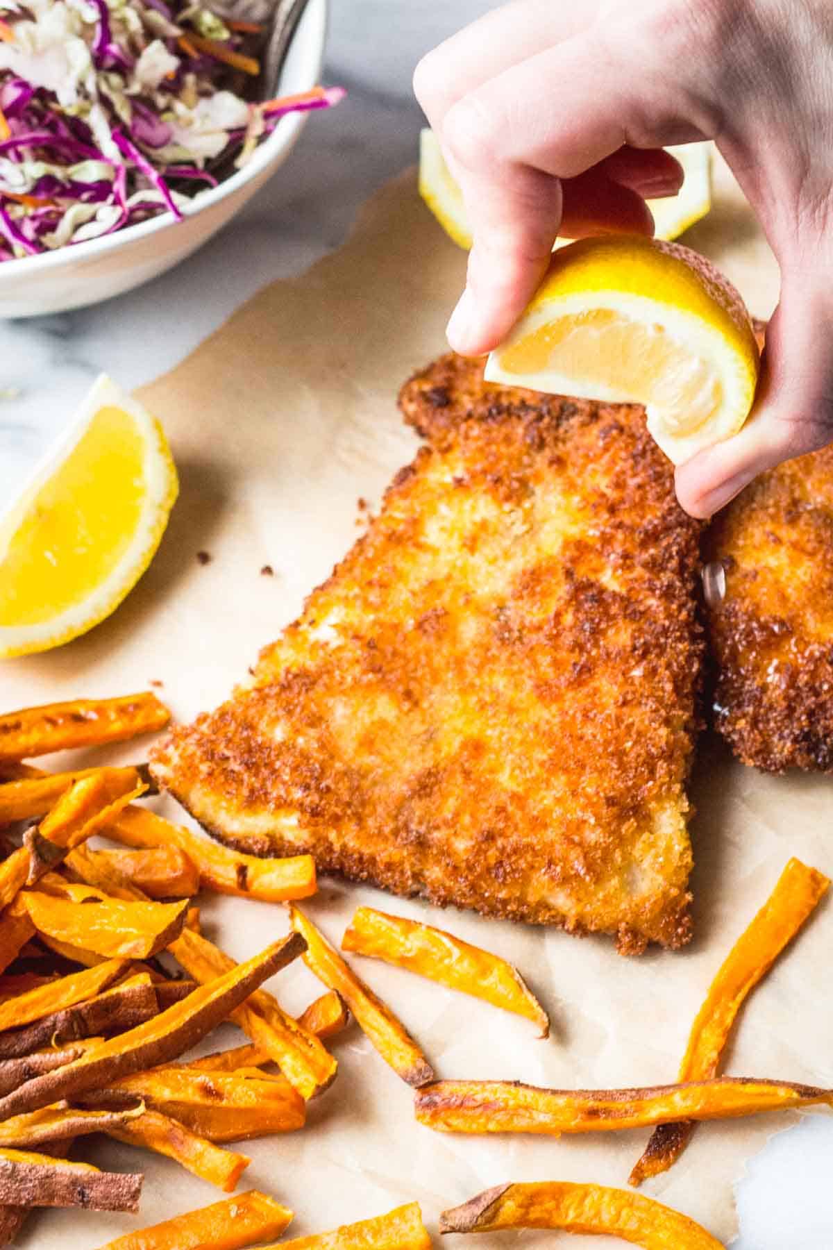A close-up action shot showing a hand squeezing a lemon wedge over a triangular piece of crispy panko-crusted fish. The golden-brown coating appears crunchy and textured. Sweet potato fries are scattered at the bottom left, and a bowl of colorful coleslaw with purple and white cabbage is visible in the upper left corner.