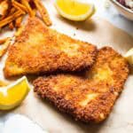 A top-down view of two triangular pieces of panko-crusted fish with a perfect golden crust, displayed on parchment paper. Four lemon wedges are strategically placed around the fish, with sweet potato fries arranged on the left side. A small portion of the coleslaw bowl is visible in the upper right corner of the frame.