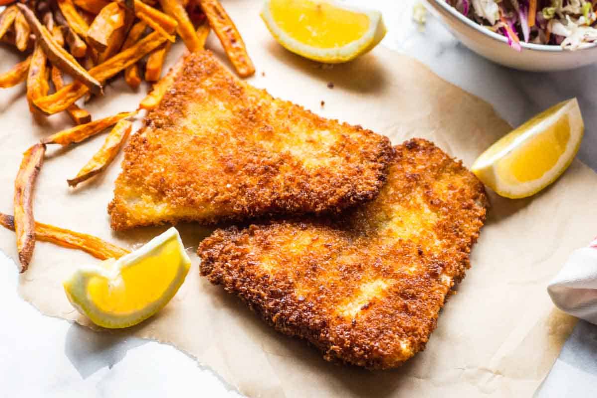 A top-down view of two triangular pieces of panko-crusted fish with a perfect golden crust, displayed on parchment paper. Four lemon wedges are strategically placed around the fish, with sweet potato fries arranged on the left side. A small portion of the coleslaw bowl is visible in the upper right corner of the frame.