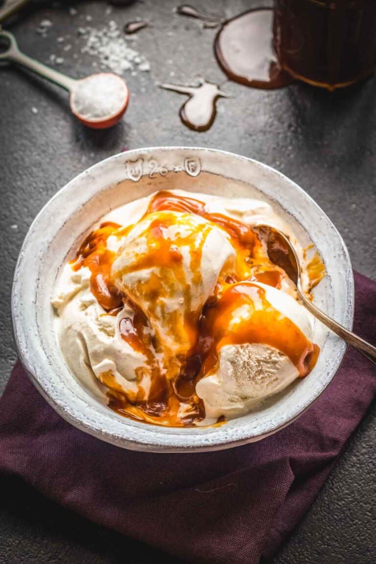 Overhead view of a bowl of vanilla ice cream topped with a thick drizzle of homemade salted caramel sauce, set on a dark surface with scattered sea salt and a jar of caramel in the background.