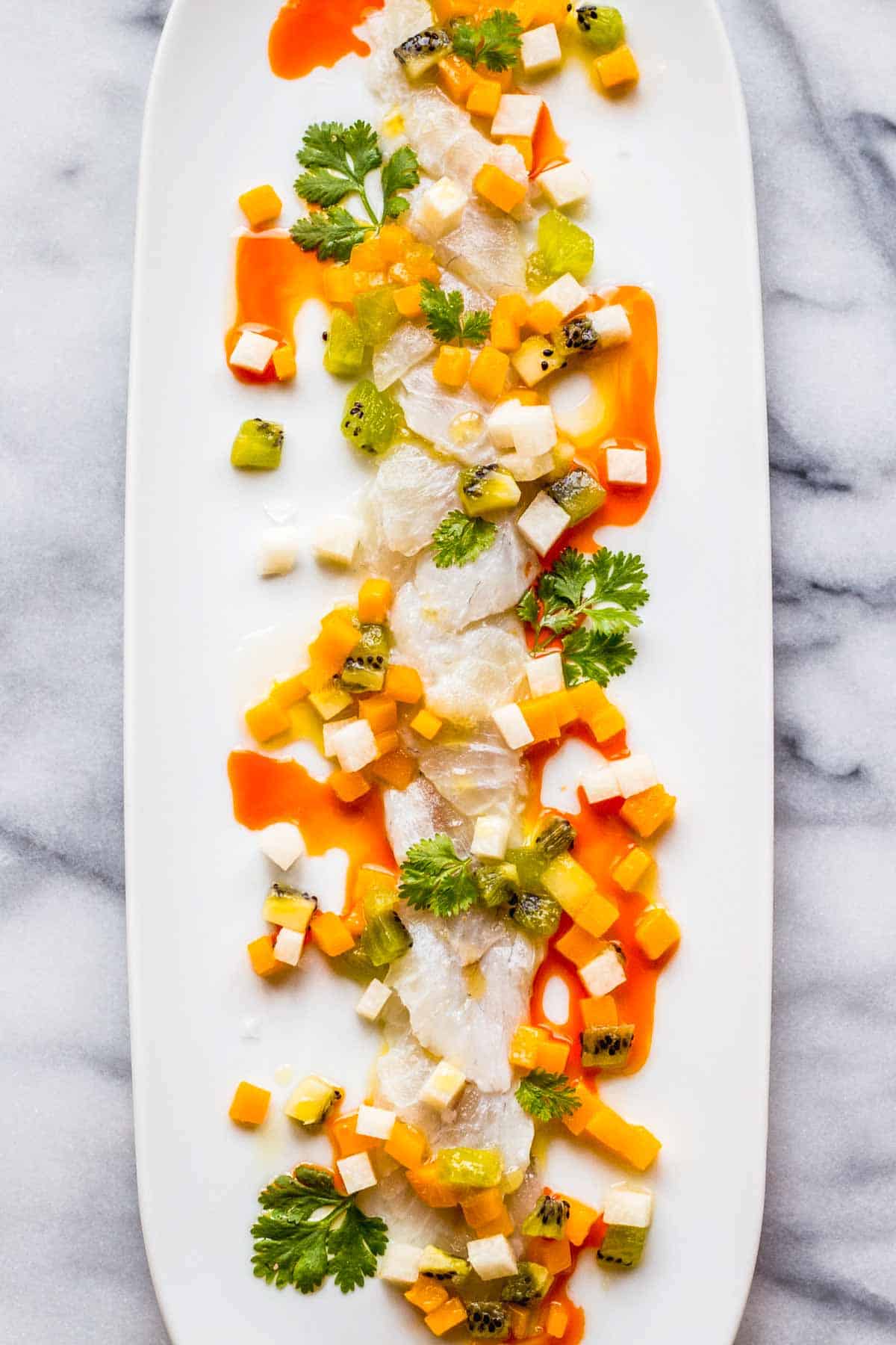 Thinly sliced raw white fish artfully arranged on a long white platter, garnished with small cubes of mango, kiwi, and jicama, drizzled with vibrant chili oil and olive oil, and accented with fresh cilantro leaves.