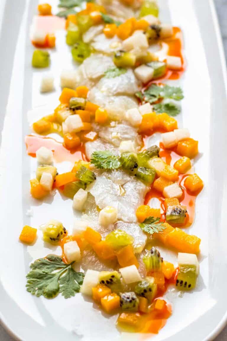 A delicate arrangement of crudo topped with colorful diced fruit and vegetables, including golden mango, green kiwi, and white jicama, with splashes of red chili oil and fresh cilantro on a white serving dish.
