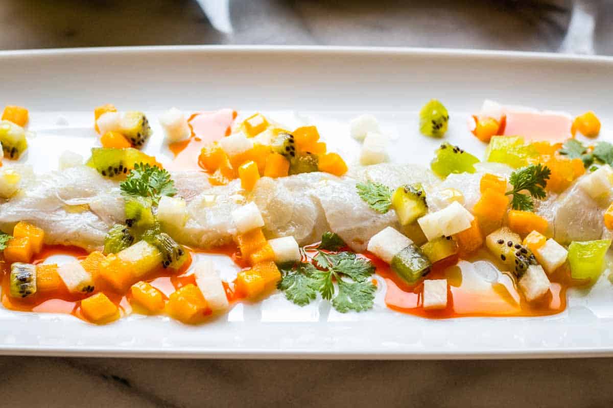 Side angle of translucent fish crudo lined along a rectangular white plate, surrounded by diced kiwi, jicama, and mango in pools of olive oil and red chili oil, with fresh cilantro leaves adding a green contrast.