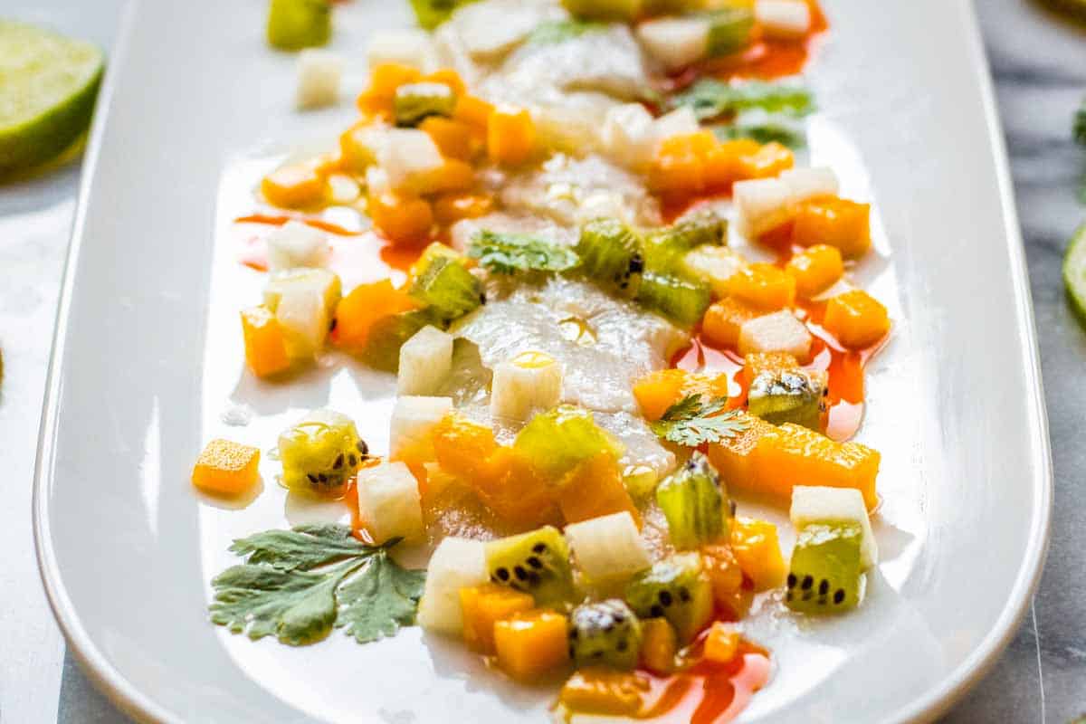 Close-up view of a plate of fish crudo with vibrant toppings of mango, kiwi, and jicama, garnished with chili oil and cilantro, creating a mosaic of textures and bright colors against the white plate.