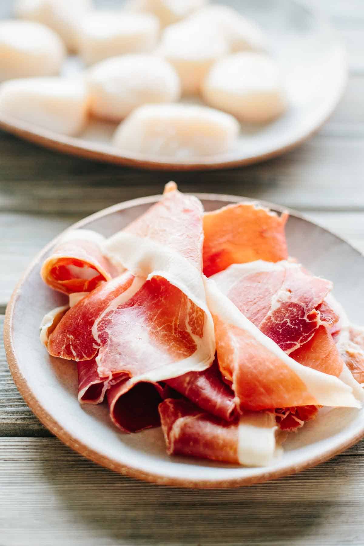 Two plates featuring the raw ingredients - a plate of thinly sliced prosciutto in the foreground and what appears to be raw scallops in the background.