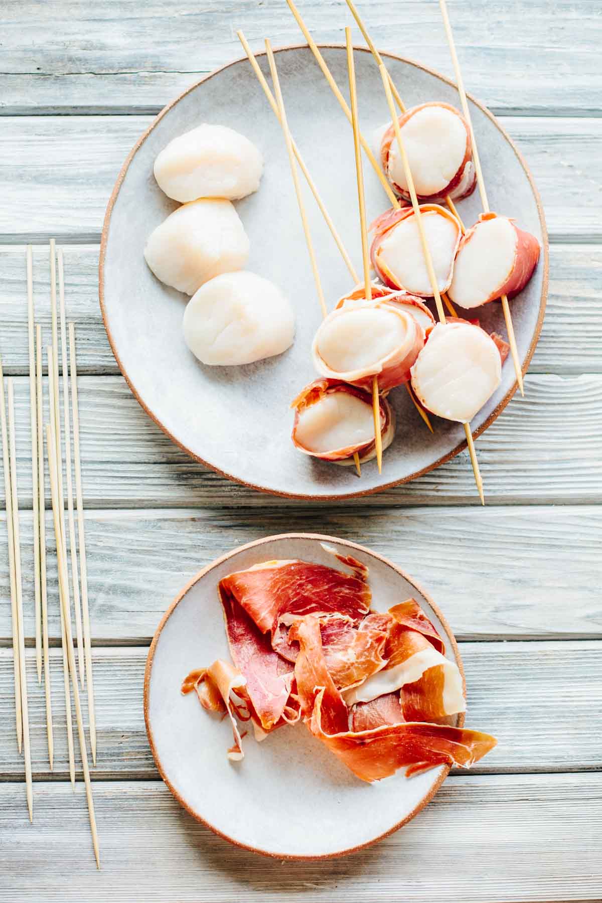 A preparation stage showing plain scallops on the left side of a plate and scallops wrapped in prosciutto on wooden skewers on the right, with additional prosciutto slices on a separate plate below.