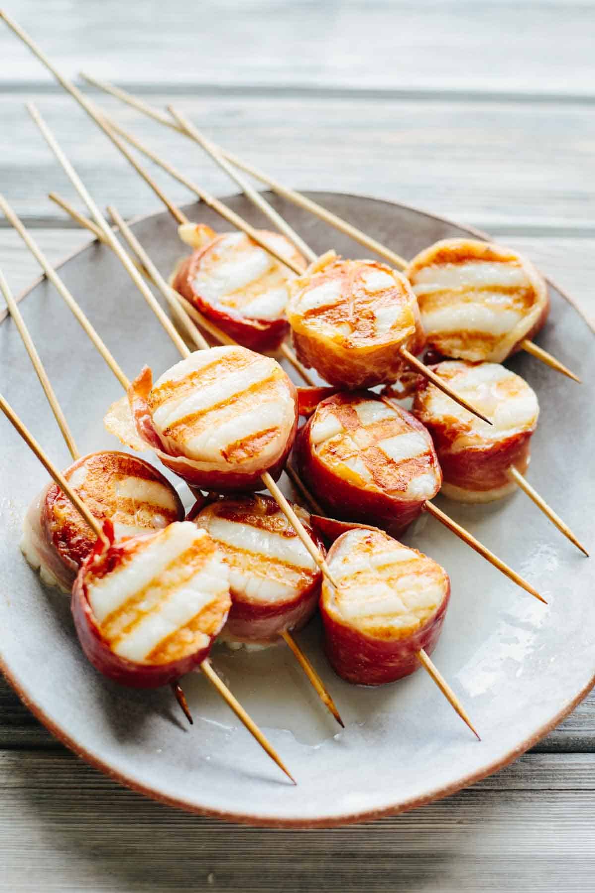 Beautifully grilled prosciutto-wrapped scallops on skewers, displaying appetizing grill marks on the scallops. The wooden skewers are arranged on a white plate.