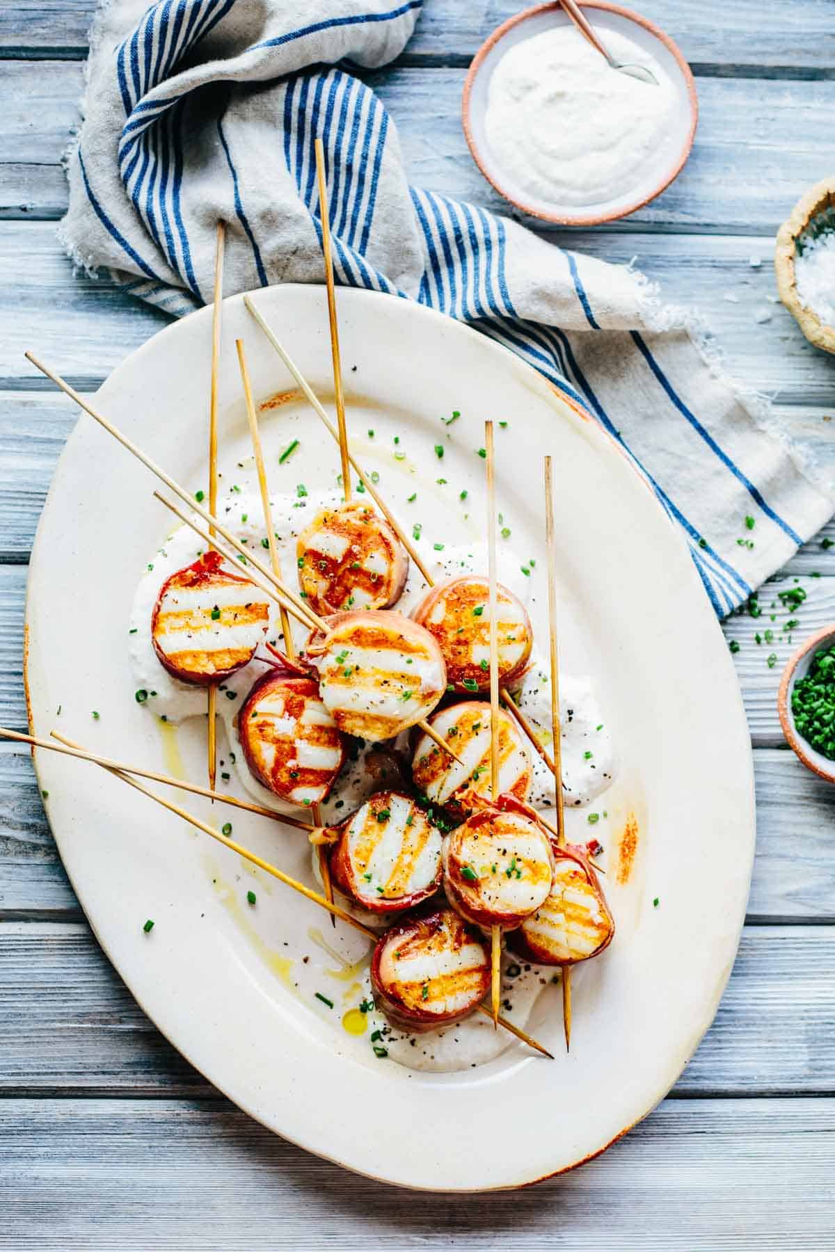 Image 4: Oval white plate featuring prosciutto-wrapped scallops on wooden skewers, served over a creamy sauce. The perfectly grilled appetizers show distinct grill marks, garnished with fresh chives. A blue-striped napkin and small bowl of sauce appear alongside.