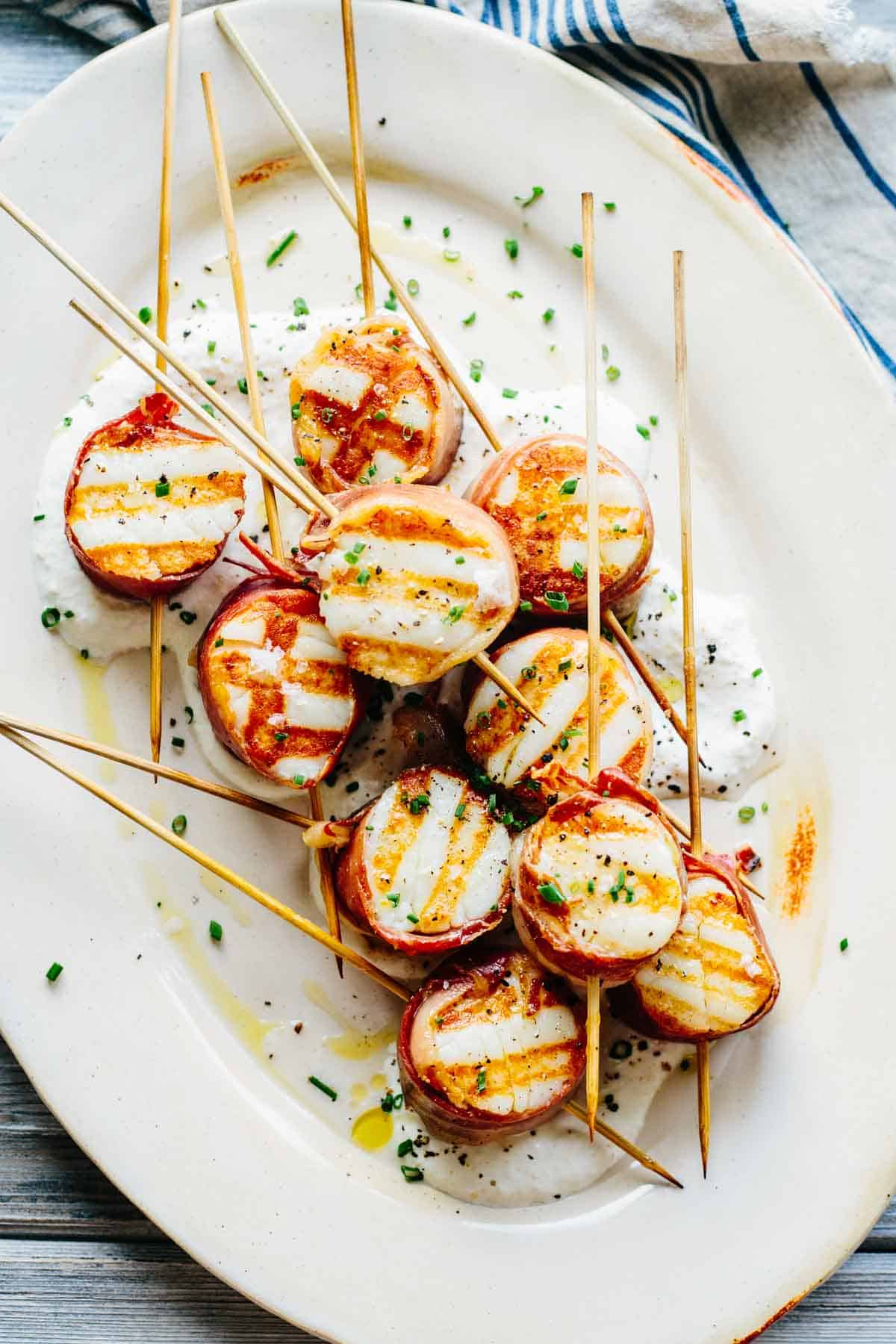 Close-up view of grilled prosciutto-wrapped scallops on wooden skewers. The caramelized scallops with crispy prosciutto are arranged on a white oval plate with a rustic rim, sitting in a pool of creamy sauce with olive oil drizzled on top.