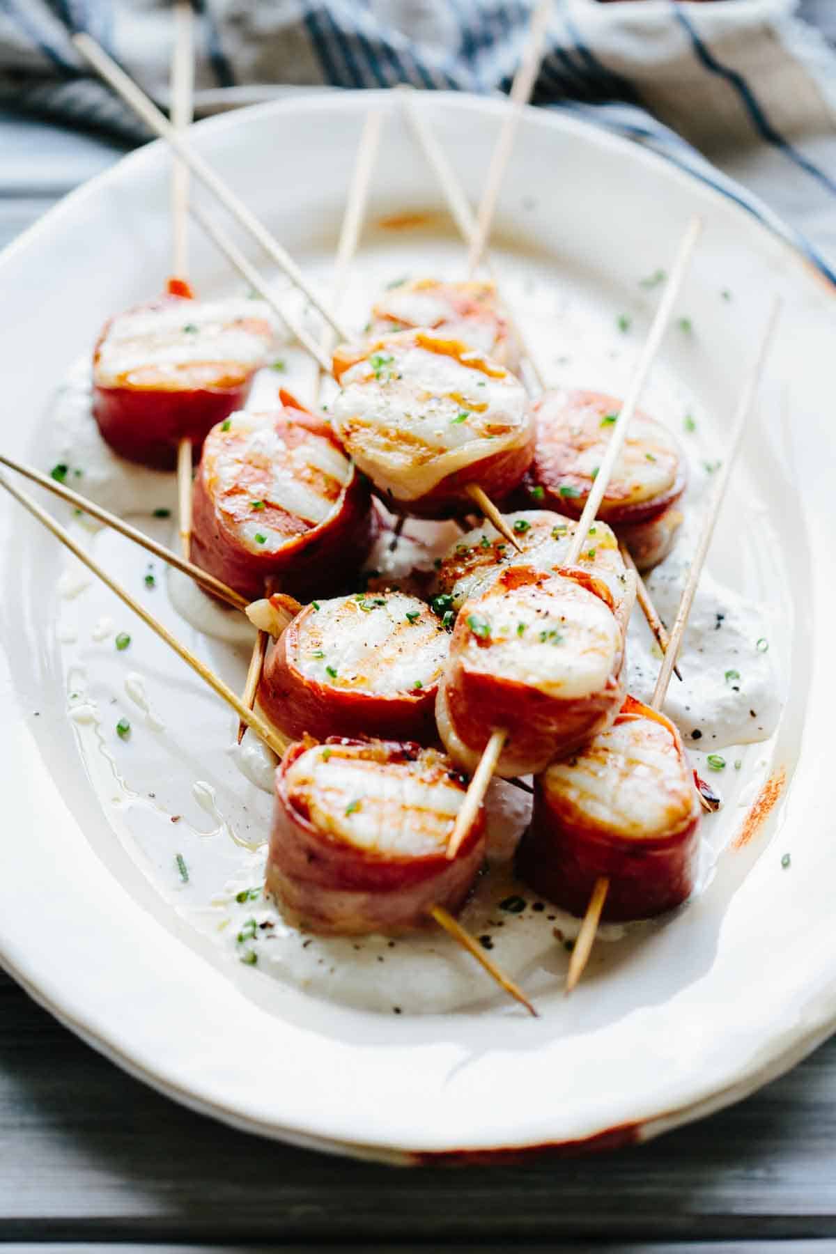 Top-down shot of prosciutto-wrapped scallops on wooden skewers fanned across a white plate. The glistening, freshly-grilled appetizers show the thin prosciutto neatly wrapped around each scallop, with a thicker creamy sauce and herb garnish beneath.