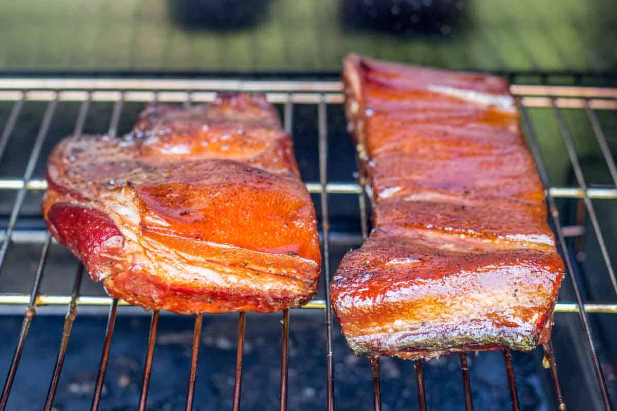 Two glistening slabs of uncooked bacon belly on a grill grate, showing vibrant reddish-pink color from curing. The meat appears moist with a glossy surface, indicating it's been freshly prepared for smoking.