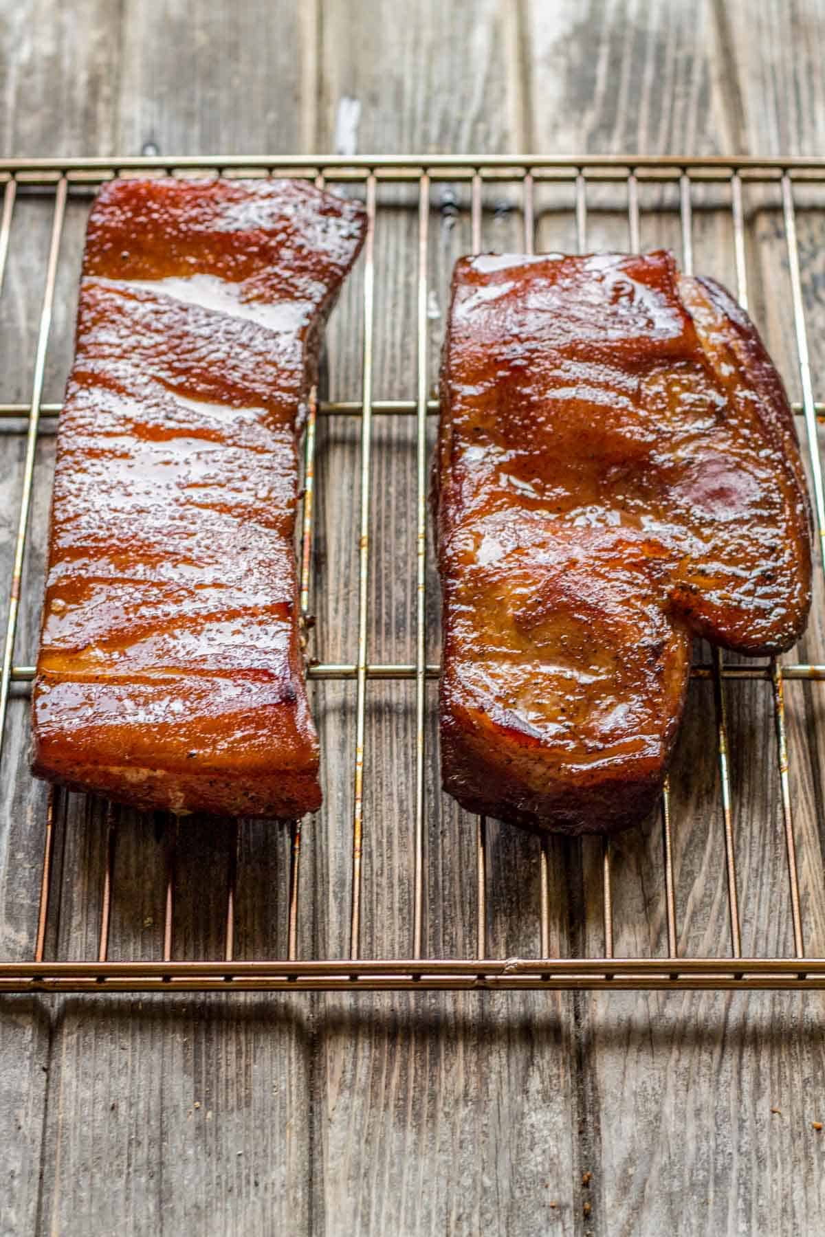 Two rectangular slabs of cured bacon resting on a cooling rack over a wooden surface. The bacon has a deep reddish-brown exterior with visible seasoning, showing the transformation after the curing process.