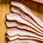 Thinly sliced bacon arranged in a fan pattern on a wooden cutting board. The slices show perfect alternating layers of white fat and pink meat with a browned exterior, demonstrating proper curing and a beautiful finished product.