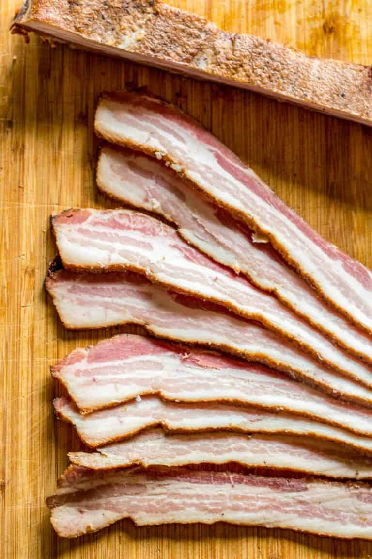 Thinly sliced bacon arranged in a fan pattern on a wooden cutting board. The slices show perfect alternating layers of white fat and pink meat with a browned exterior, demonstrating proper curing and a beautiful finished product.