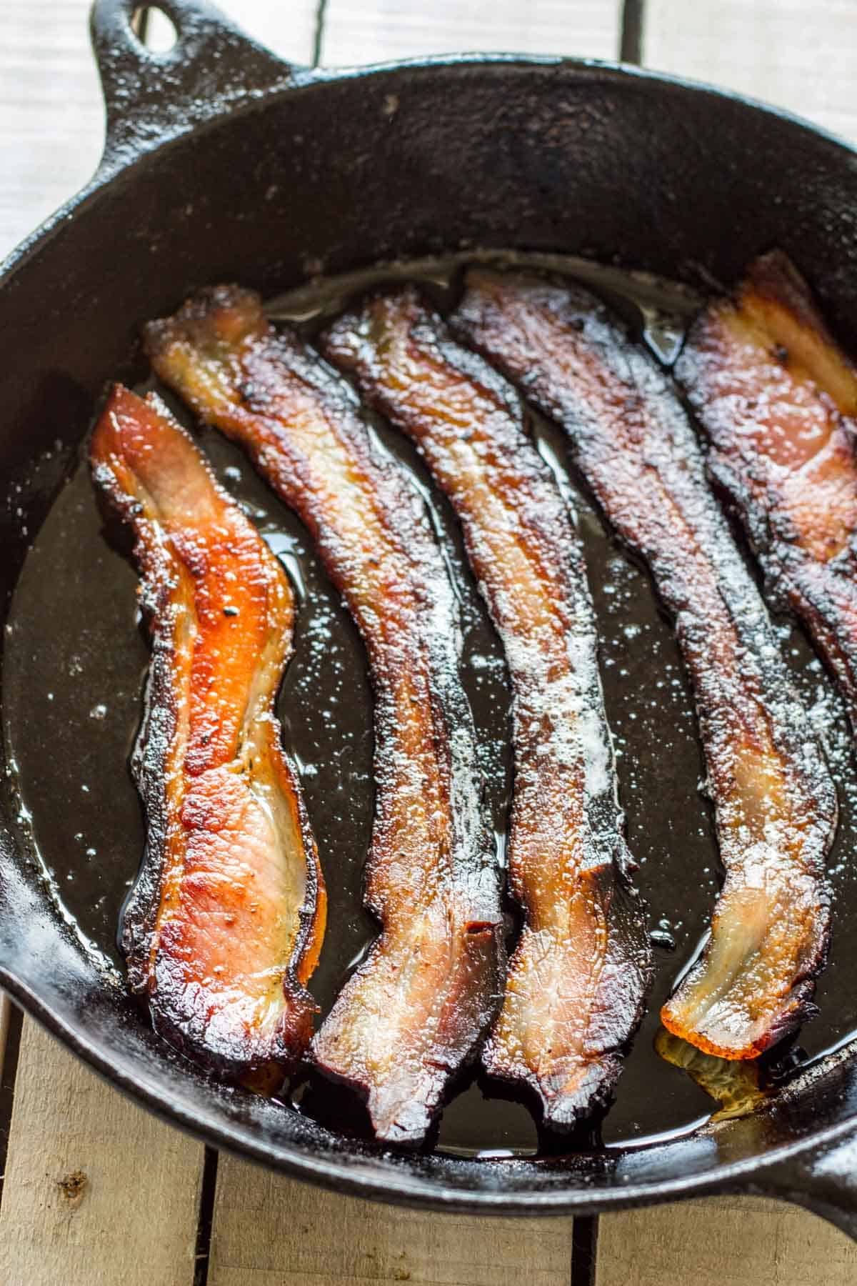 Strips of freshly cooked bacon sizzling in a cast iron skillet with rendered fat. The bacon strips show crisp edges with caramelized surfaces.