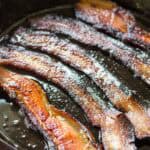 Bacon strips cooking in a cast iron pan with bubbling fat. The strips have achieved a deeper caramelization with more pronounced crisping around the edges and glistening surfaces.