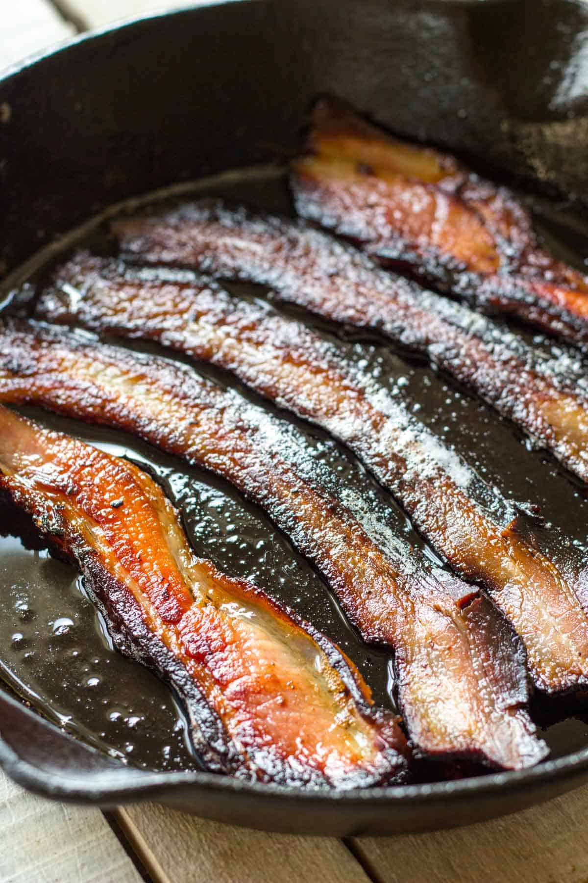 Bacon strips cooking in a cast iron pan with bubbling fat. The strips have achieved a deeper caramelization with more pronounced crisping around the edges and glistening surfaces.